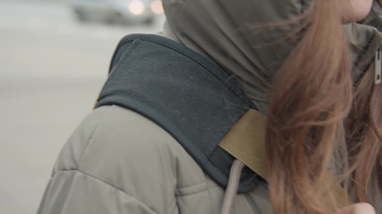 Young woman adjusting backpack strap on shoulder, closeup of coat and brown hair, zipper detail and fabric texture, morning commuter on urban sidewalk, candid routine and quiet movement