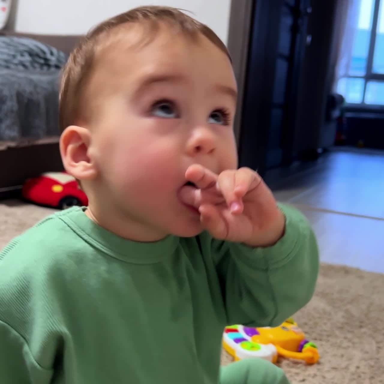 Little kid sits on the floor near his toys. Baby boy looks up with interest taking a finger into mouth. Close up side view