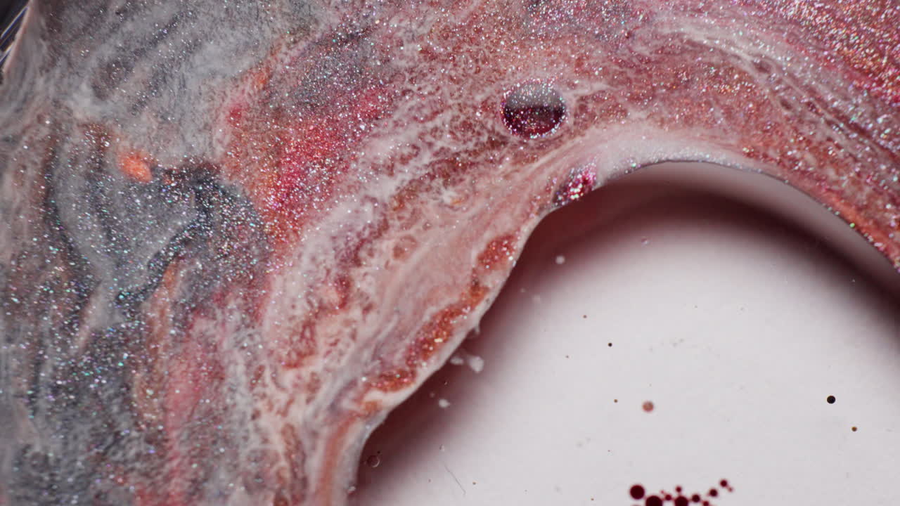 Macro shot of glittery grey and pink inks disturbed by a sudden blast of milky liquid, causing shimmering pigments to scatter