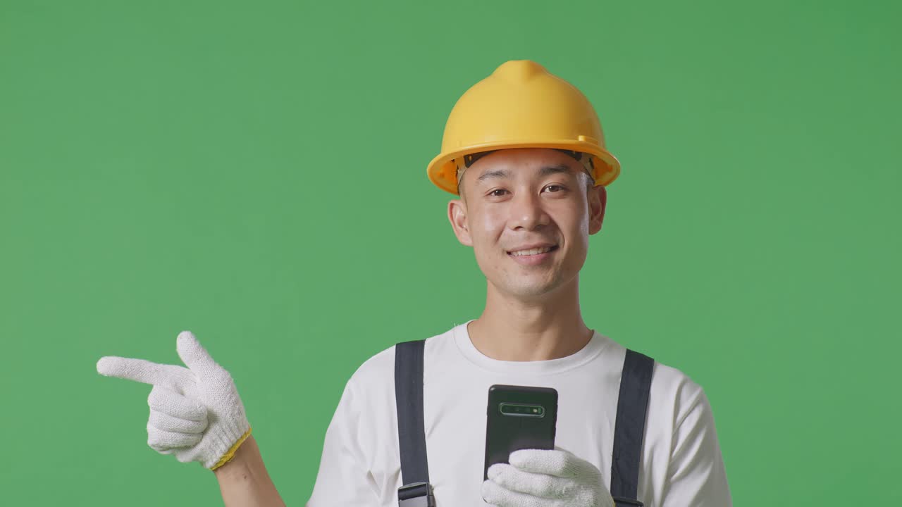 Close Up Of Asian Man Painter Wearing Safety Helmet Using Smartphone And Pointing To Side While Standing In The Green Screen Background Studio