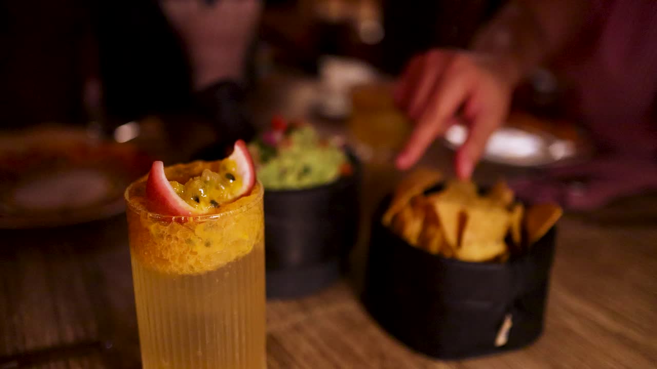 Una mano prende patatine di mais accanto a guacamole e un cocktail ghiacciato al frutto della passione su un tavolo di legno, in un'illuminazione calda e d'ambiente di un ristorante. Bassa profondità di campo, primo piano.