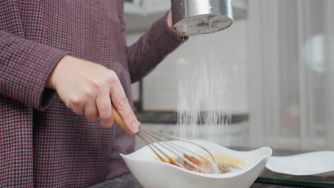 cook stirring mixture in white bowl while sifting wheat flour with stainless steel sieve, flour falls gently into bowl as ingredients mix, showcasing smooth texture, kitchen setting with modern countertop