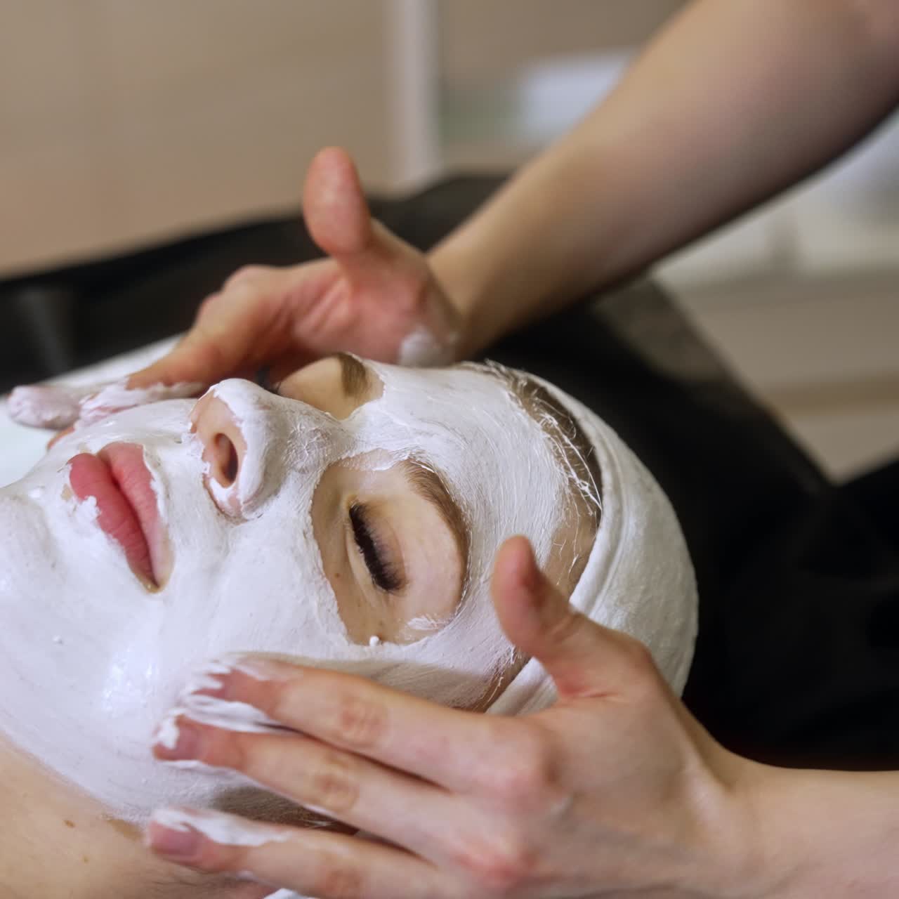 Gentle movements of cosmetologist's hands on the client's face. Woman lies eyes closed having facial mask applied on