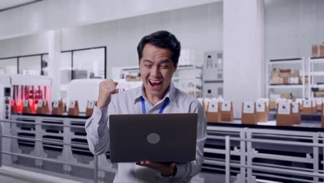 asian male professional worker standing with his laptop with conveyor for packaging coffee beans in bags at coffee factory, he raises his fist up with screaming goal