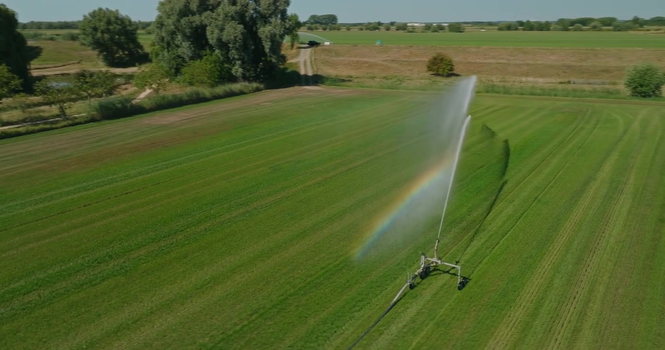 Orbita aerea di un drone attorno a un grande irrigatore agricolo che irriga un terreno agricolo olandese in una giornata di sole, creando un vivido arcobaleno e una bellissima ombra d'acqua attraverso il campo.