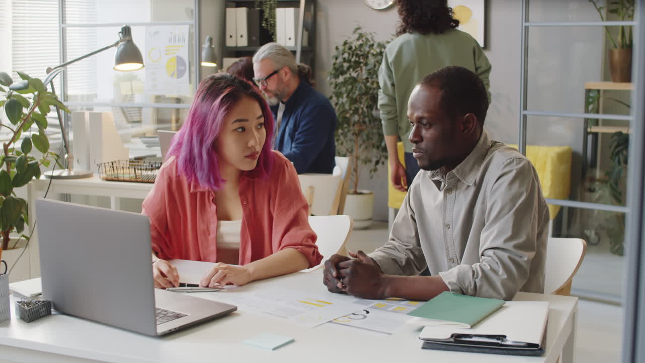Multiethnic Mentor and Trainee Talking and Using Laptop in Office