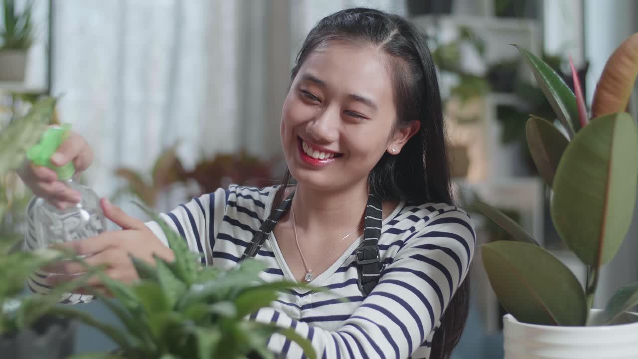미소 짓는 아시아 여성이 집에서 식물에 물을 주고 있는 장면