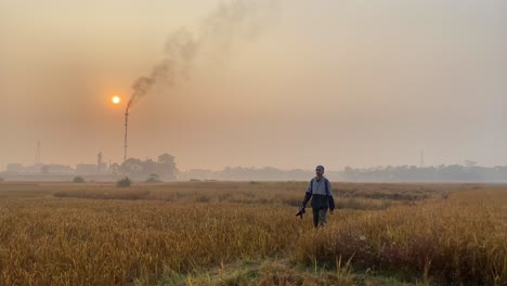 photographer walking by camera in vast paddy field with gas burning plant behind