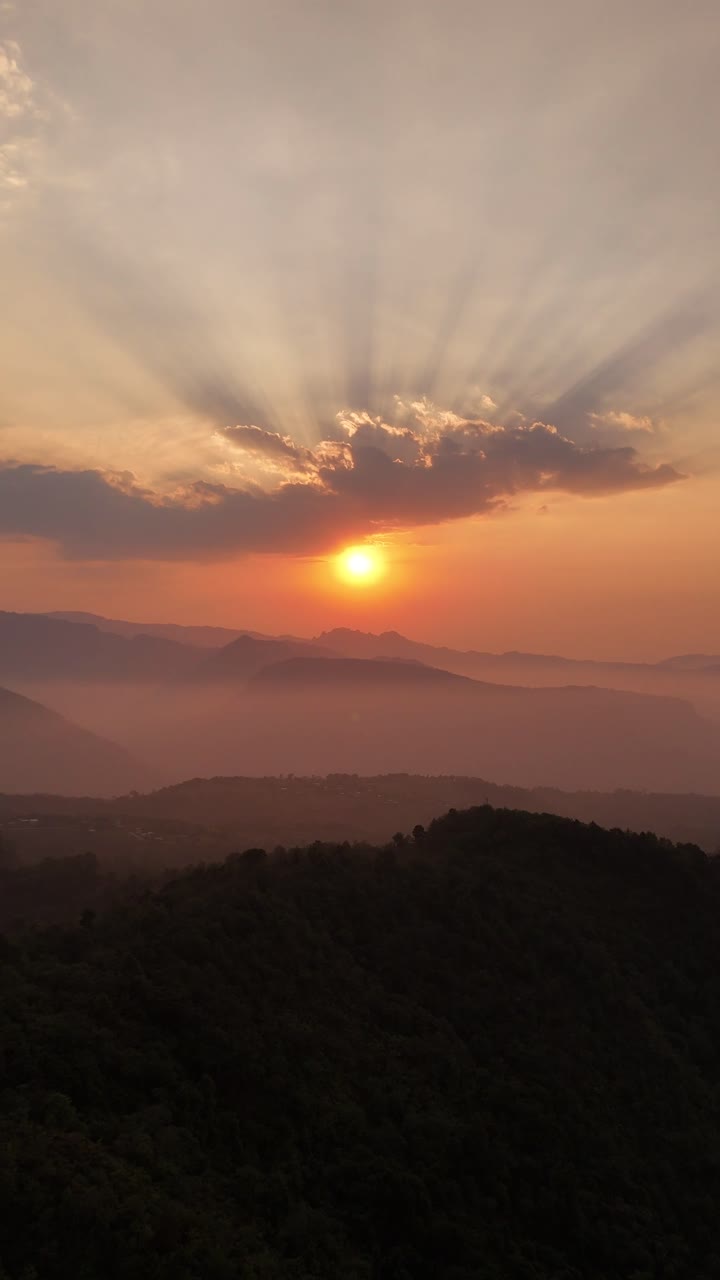 A vertical view of a red sky at sunset over mountains, with sun rays breaking through clouds.