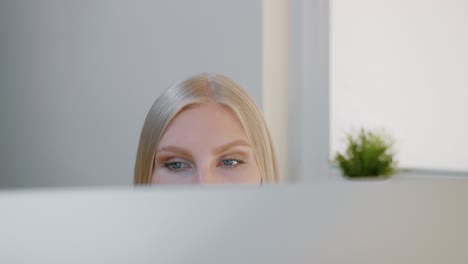 female looking at computer monitor. blond attractive woman sitting at window and looking attentively at computer screen shot made over top of monitor.