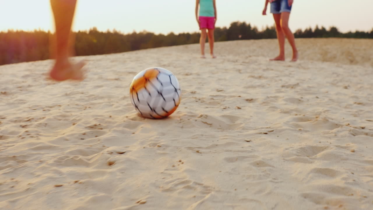 Familia jugando al fútbol en el encuadre de las piernas del hombre golpeando el balón. El balón en la arena.