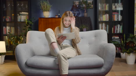 Mujer-Joven-Feliz-Con-Auriculares-Y-Tableta-Entrando-En-La-Sala-De-Estar-Y-Sentada-En-El-Sofá