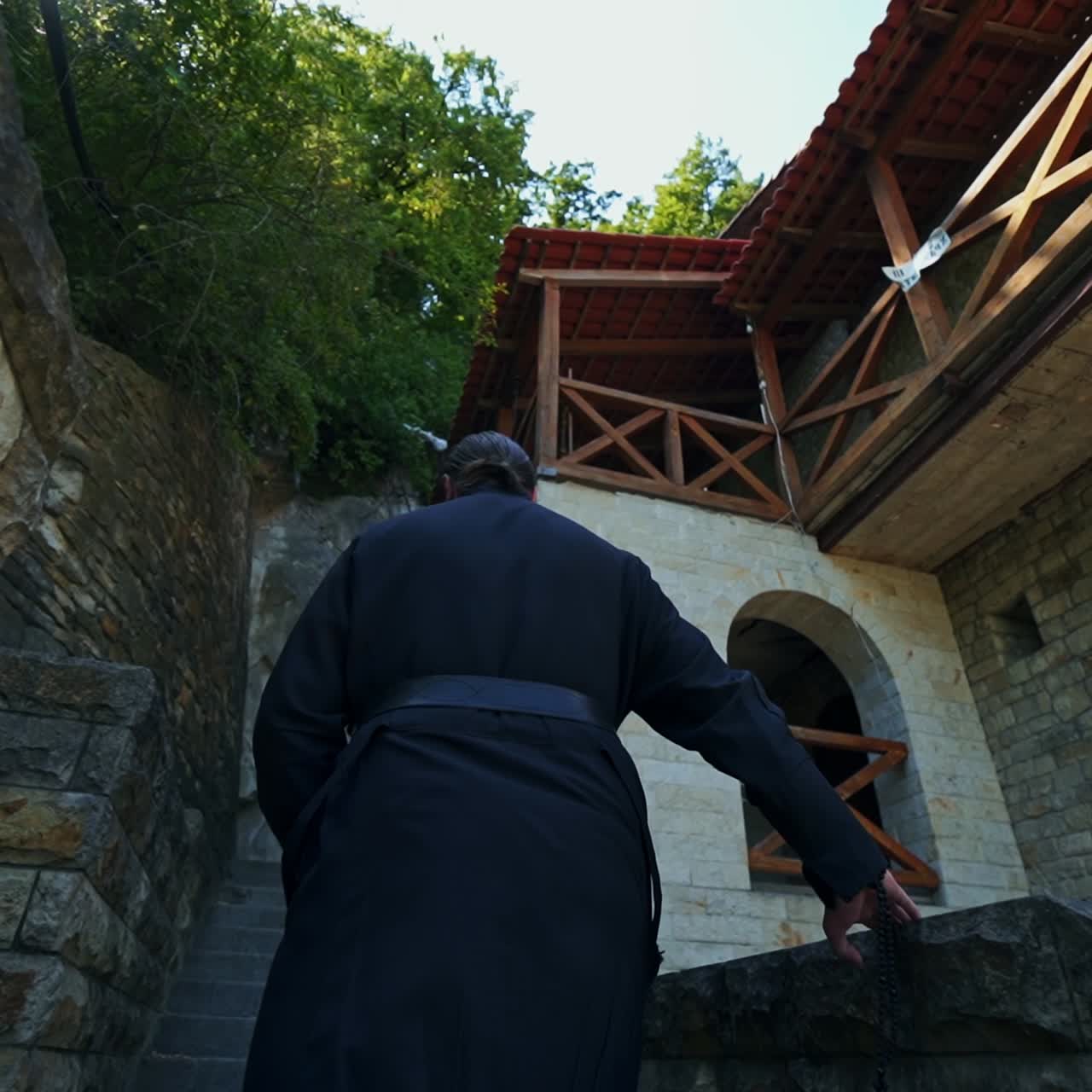 Following the priest wearing black robe. Orthodox monk going up the stone stairs of the monastery. Low angle view