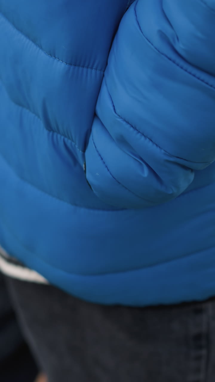Close-up of an individual's arm and side, wearing a bright blue puffer jacket and holding a shopping bag, with blurred urban background featuring greenery by the wayside