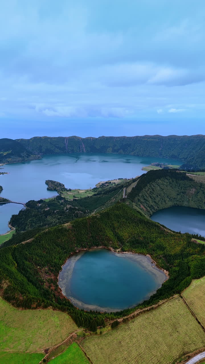 Craters of sleeping volcanoes filled with lakes. Picturesque village is located on the waterfront. Panorama of the Azorean Islands. Top view. Vertical video.
