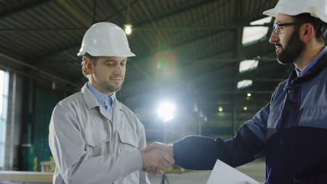 two engineers wearing helmets and holding blueprint while talking and shaking hands in a factory