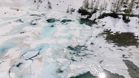 Vista-Aérea-De-Un-Lago-Congelado-Parcialmente-Cubierto-De-Hielo-Agrietado,-Rodeado-De-Suelo-Y-árboles-Cargados-De-Nieve,-Que-Ilustra-La-Belleza-De-La-Naturaleza-Invernal.