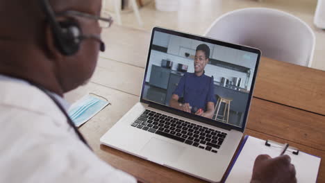 african american male doctor wearing phone headset taking notes while having a video call on laptop