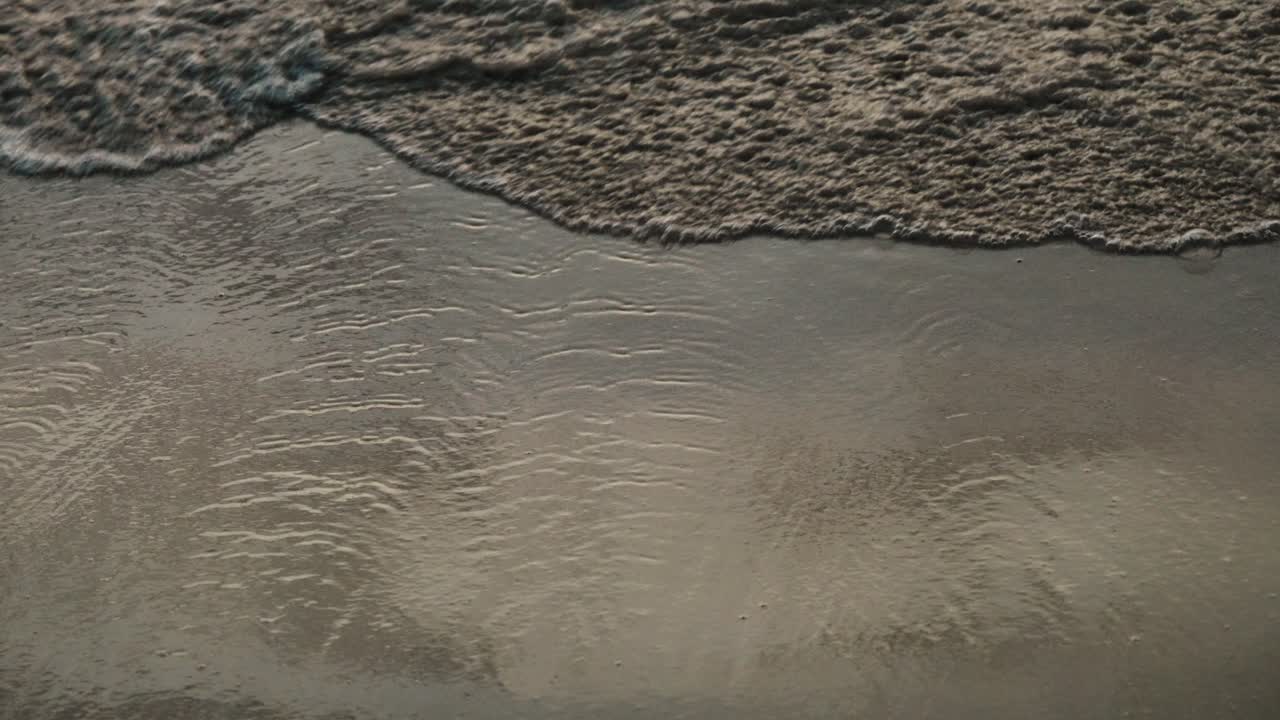 A soft wave gently washes over wet shoreline sand under warm evening light