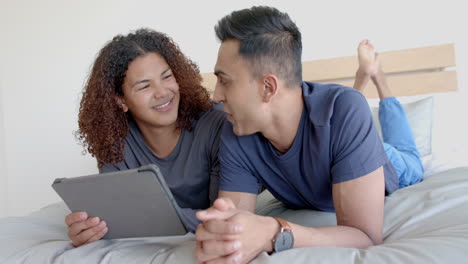 Happy-diverse-gay-male-couple-lying-on-bed-using-tablet-and-talking-in-the-morning,-slow-motion