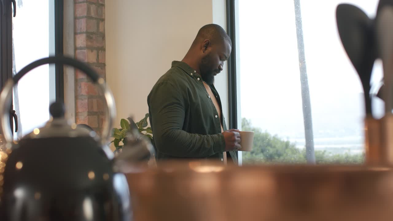 Homem afro-americano de meia-idade olhando pela janela, depois inspecionando a caneca, preparando-se para tomar um gole na cozinha