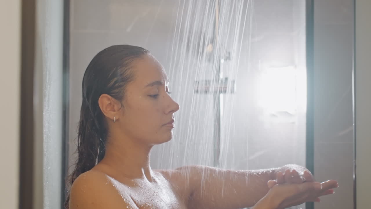 Hotel shower woman gathers warm water in hands under falling stream, side profile in soft steam, calm bathing ritual with gentle light and toiletries visible, reflective relaxation scene