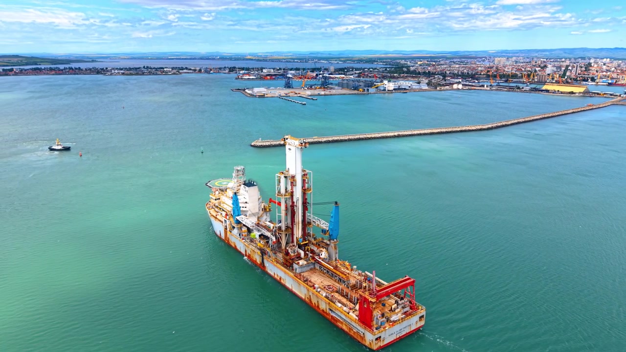 Luftfoto af stort industrielt boreskib i byens havnebugt. Droneoptagelse i fugleperspektiv af et offshore boreskib med tårn og kraner nær havnekajer og en bølgebryder.