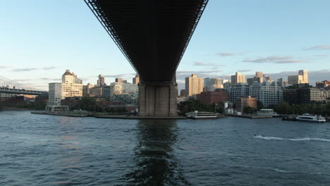 wide angle aerial tracking shot underneath the brooklyn bridge with jet skis passing by and brooklyn in the background at dusk on a clear summer evening