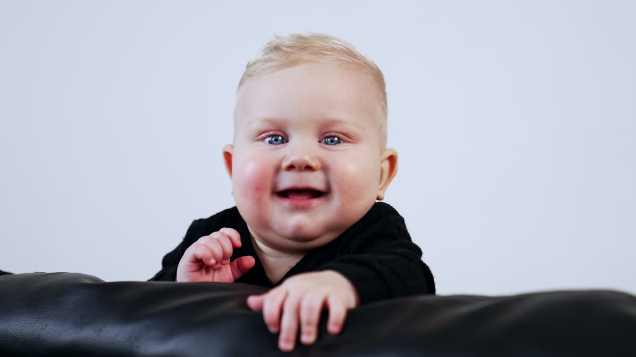 Smiling chubby baby boy in black sweater on the leather sofa. Cute child portrait at white backdrop.