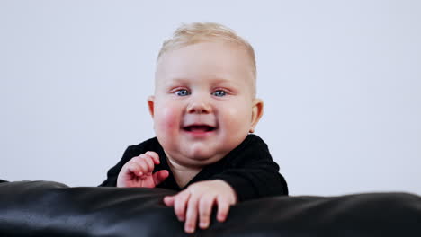 smiling chubby baby boy in black sweater on the leather sofa. cute child portrait at white backdrop.