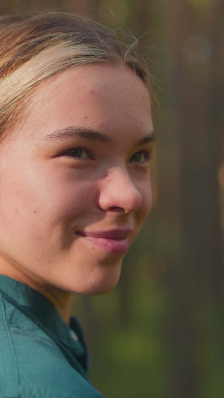 primer plano de una mujer con camisa verde de pie al aire libre en el bosque, girando la cabeza con una sonrisa sutil mientras sopla un beso lúdico en el aire, la luz del sol se filtra a través de los árboles