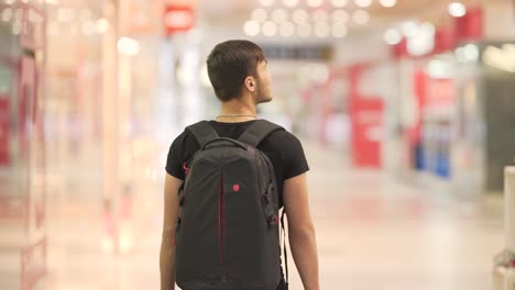 chico joven con una mochila negra caminando en el centro comercial y mirando a través de las tiendas