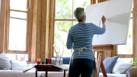 Senior-biracial-woman-wearing-apron-and-painting-on-big-canvas-at-home,-slow-motion