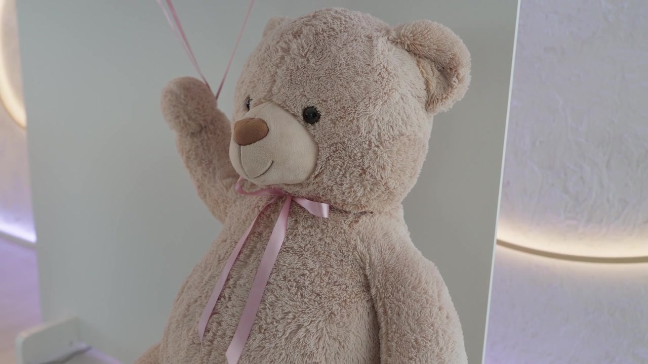 Boneka Beruang Teddy Mewah Besar Berwarna Krem Mengenakan Pita Merah Muda di Dalam Tempat Acara yang Terang.