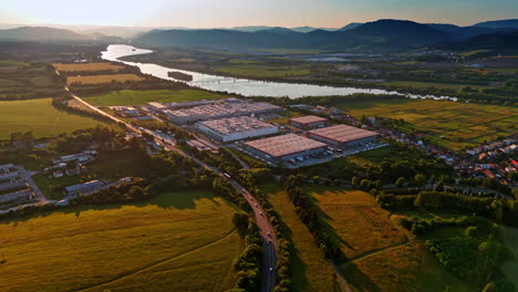 industrial area near river at sunset. large industrial complex along riverbanks with surrounding fields and mountains during golden hour in the evening