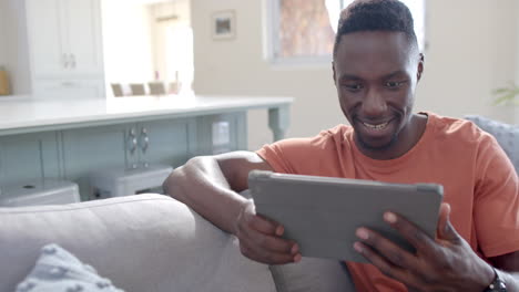 Happy african american man sitting and using tablet in sunny living room, slow motion