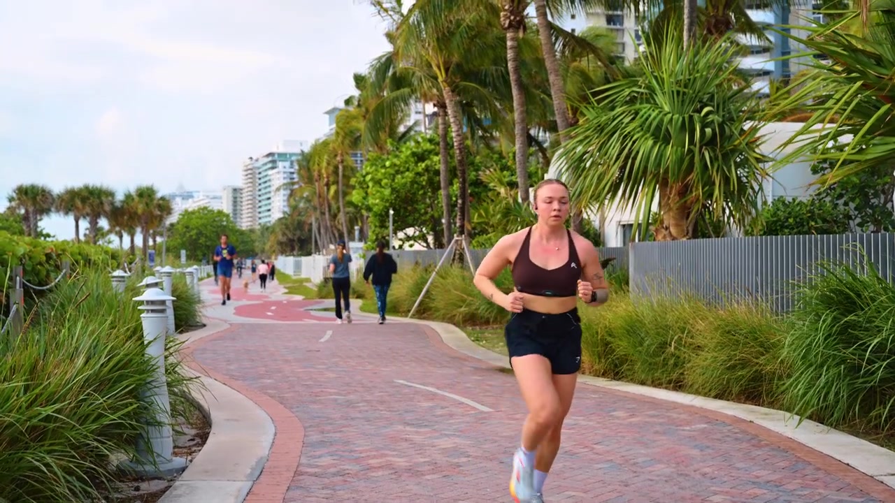 Miami, USA, 16 aprile 2026: Donna che fa jogging su una passeggiata pavimentata sulla spiaggia, fiancheggiata da palme. Vista frontale di una donna che corre su una promenade in mattoni circondata da palme, vegetazione tropicale e passanti.
