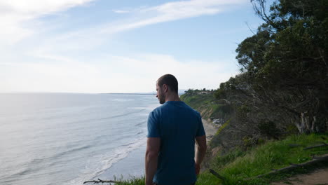 Un-Joven-En-Forma-Caminando-Por-El-Borde-De-Un-Acantilado-Oceánico-Mirando-Hacia-La-Playa-En-Un-Día-Soleado-En-La-Naturaleza-Durante-Una-Caminata-En-Santa-Barbara,-California-A-Cámara-Lenta