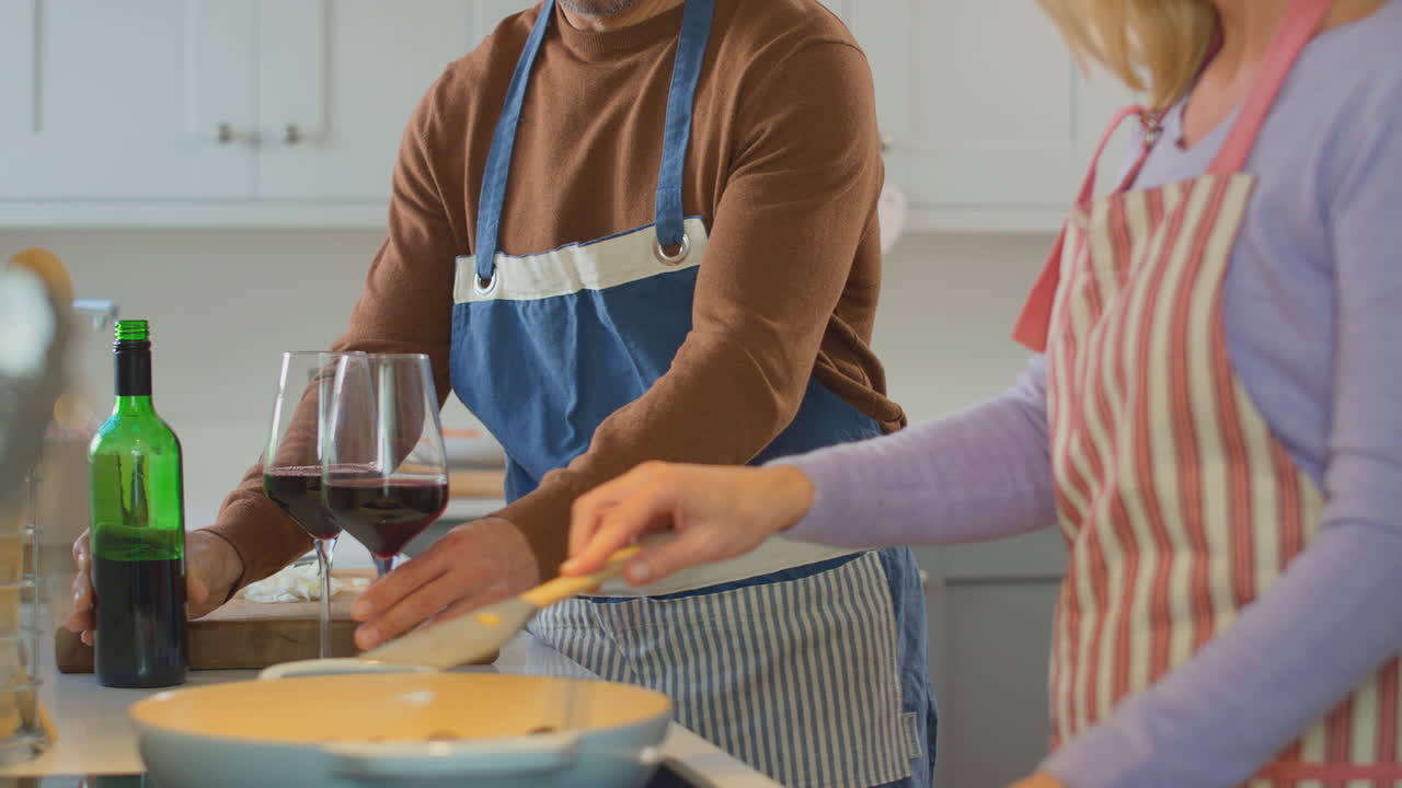 Coppia matura che indossa grembiuli, beve vino rosso mentre prepara gli ingredienti e cucina un pasto a casa insieme - ripreso al rallentatore