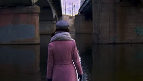 woman walking under a bridge by a river in pink coat