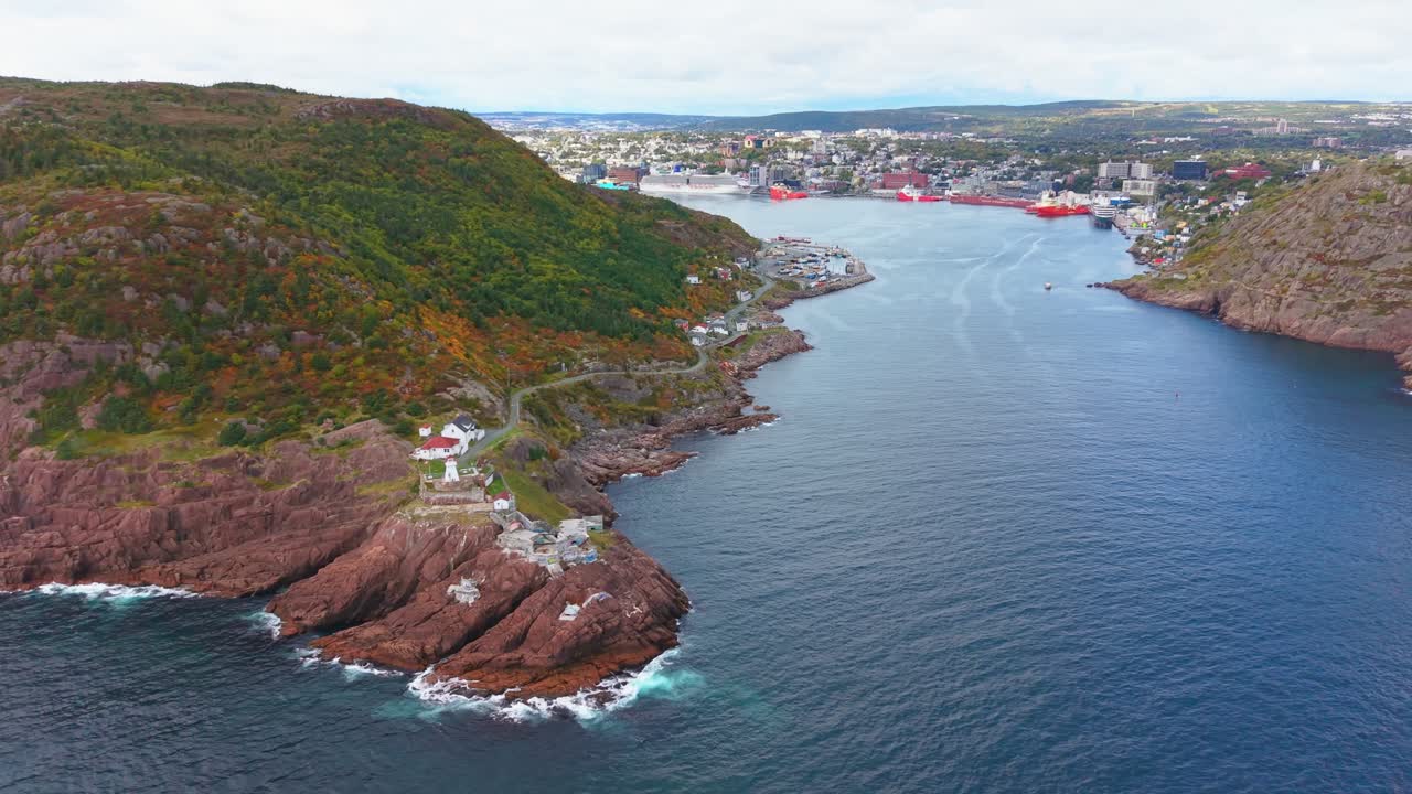A high-altitude drone flight over Fort Amherst reveals red escarpments and turquoise waters, capturing autumn foliage on hills and cottages nestled around a calm bay under clear skies