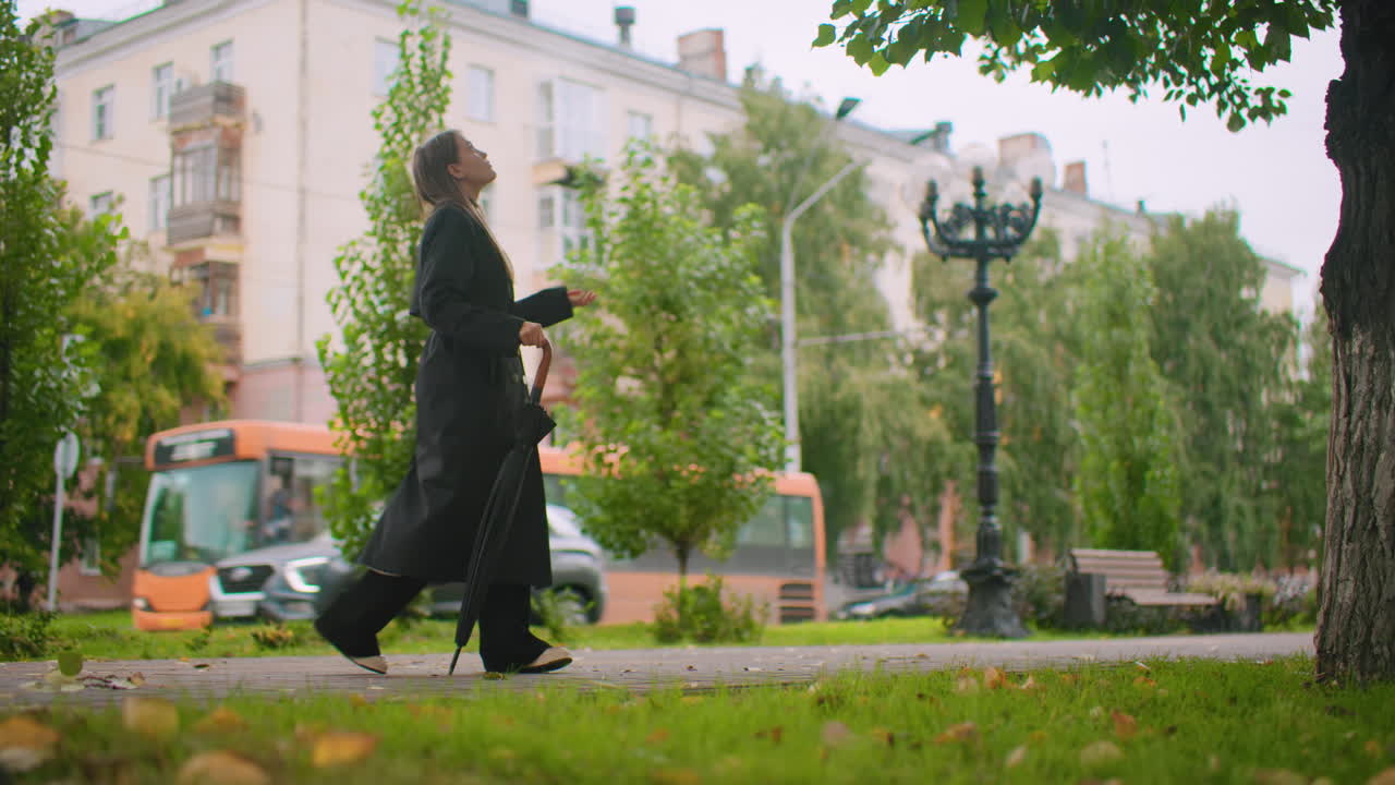 Woman in long black trench coat walking on park sidewalk holding closed umbrella, autumn leaves scattered on ground, city background with buildings, trees, lamp post, passing bus weather mood