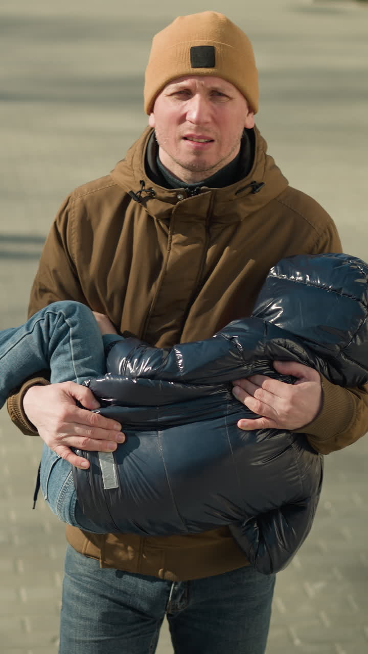 Father carrying his sleeping child while walking, visibly stressed, the father is wearing a brown jacket and beanie, and the child is in black jacket, shadows are visible