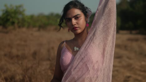 woman in pink sari standing in dry field, intense gaze, sunlit backdrop, close-up