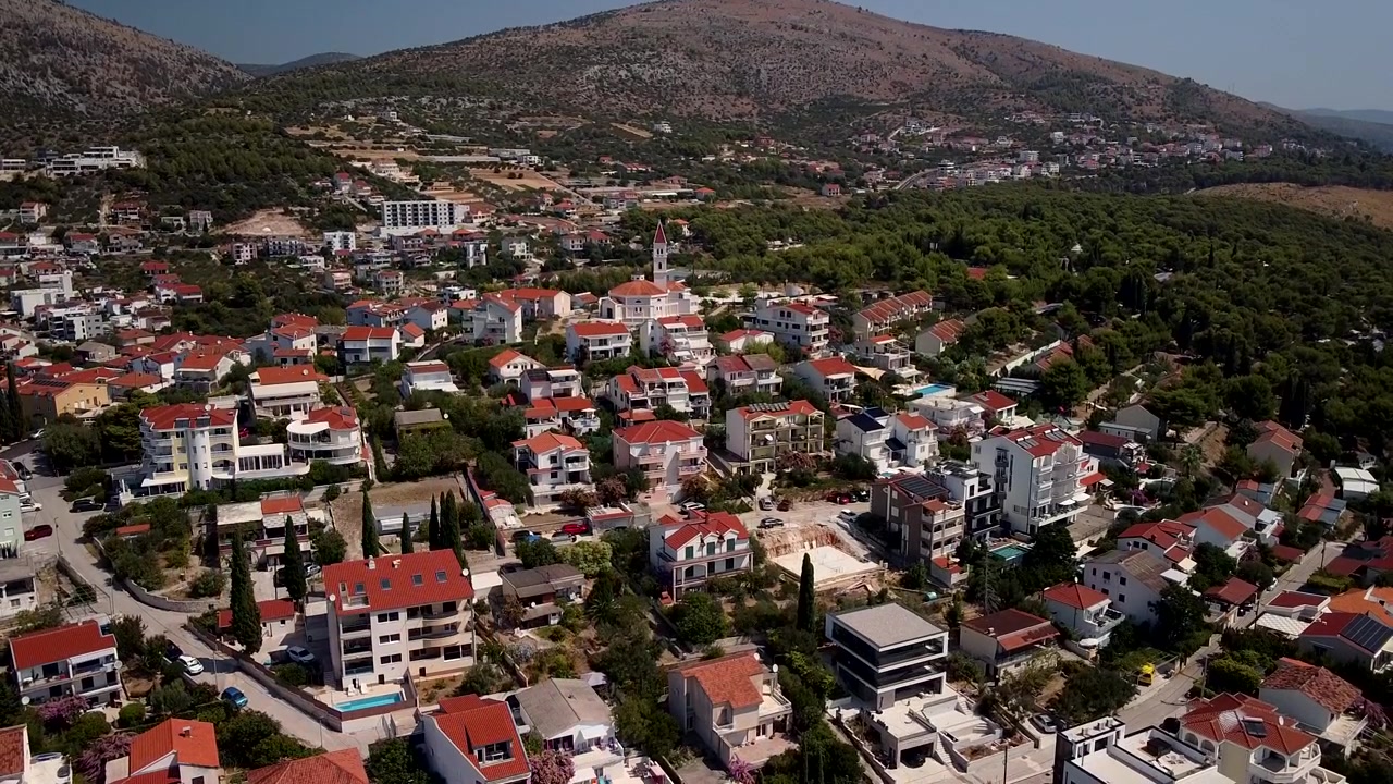 Seget Vranjica, Trogir, Chorvatsko – domy s červenými střechami rozprostřené po pobřežním svahu, letecký záběr stoupající a s náklonem dolů přes kostely a oranžové domy.