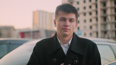 business mogul standing close to luxury car lost in thought with hand on suit collar, background features modern office building and parked cars in city business district