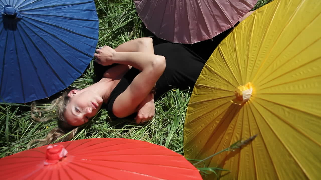 한 여자가 나의 다양한 색깔의 우산으로 둘러싸인 풀밭에 등을 대고 누워 있습니다.
