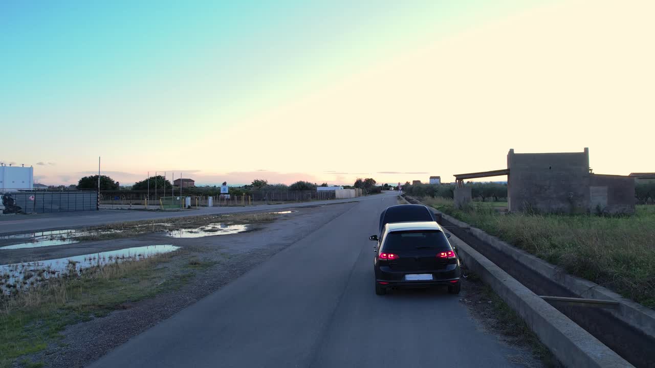 Vista aerea di un'auto in panne parcheggiata su una strada rurale, con un lampeggiante arancione V16 di emergenza sul cofano aperto