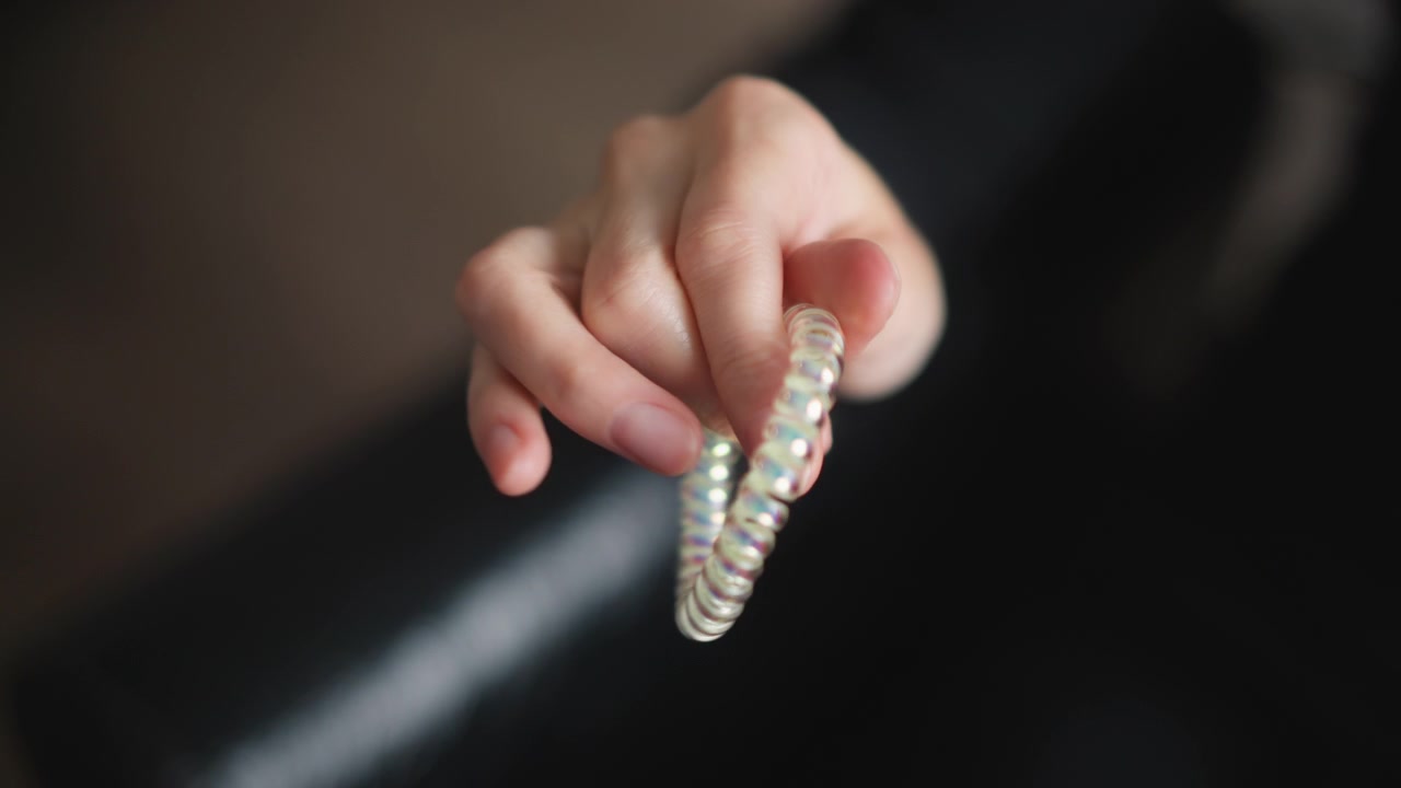 Primo piano di una mano che tiene e manipola un elastico per capelli a spirale iridescente, dimostrandone la flessibilità. L'oggetto riflette la luce con sottili sfumature arcobaleno mentre viene allungato tra le dita. Luce soffusa.