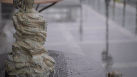 Escultura-Decorativa-De-Piedra-De-Un-Niño-Sosteniendo-Un-Pez-Encima-De-Un-Pedestal-De-Granito-En-Un-Día-Lluvioso